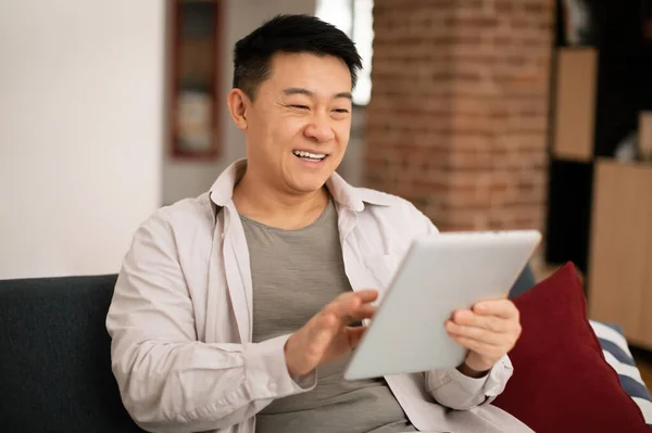 在家里的沙发上 穿着休闲装的快乐的亚洲成熟男人 使用数码平板电脑和微笑 在社交媒体上滚动 复制空间 — 图库照片