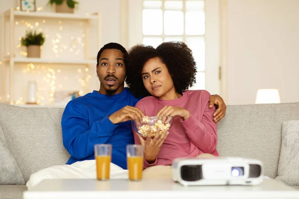 国内エンターテインメント 不気味な映画を見てショックを受けたアフリカ系アメリカ人の配偶者はポップコーンを食べ 自宅でプロジェクターの近くに一緒に座っています デジタルレジャーと楽しいコンセプト 選択的フォーカス — ストック写真