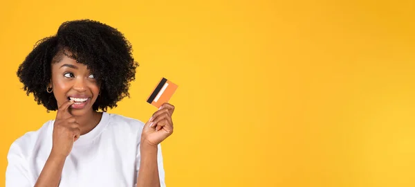 幸せな若いアフリカ系アメリカ人の女性を購入することを夢見て クレジットカードを見て 考えて 黄色のスタジオの背景に隔離された オンラインショッピング 現金のためのショッピング — ストック写真