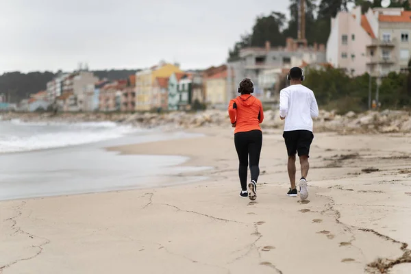 穿着运动服和无线耳机的年轻黑人夫妇在海滩上跑步 享受夏天的户外活动 身体护理和人际关系 共同运动和自然 — 图库照片
