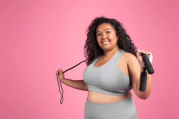 Satisfied black woman plus size in sportswear with skipping rope ready to training on pink background, smiling at camera, free space. Cardio active workout, weight loss and fitness