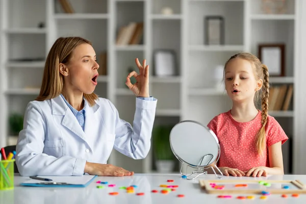 Speech Therapy Concept. Cute little girl pronouncing sound O looking at mirror, professional therapist lady teaching female child right pronounciation during private lesson at office