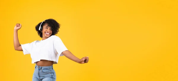 ワイヤレスヘッドフォンダンスで幸せな若いアフリカ系アメリカ人の巻き女性は 黄色のスタジオの背景 パノラマに隔離された楽しさを持っています 女性は音楽やオーディオアプリを楽しむ — ストック写真