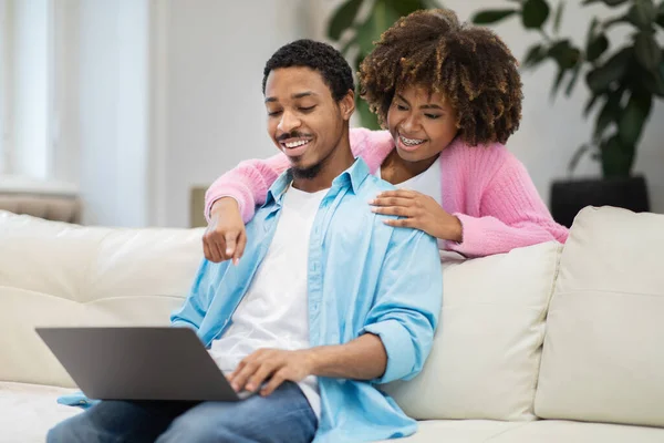 Cheerful young black spouses sitting on couch, planning vacation, using laptop at home. Pretty african american lady hugging her husband from behind and pointing at computer screen, copy space