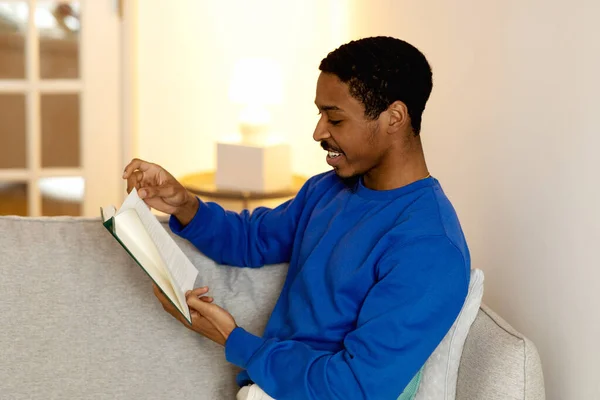 Literature Enjoyment. Side View Shot Of Happy Black Man Reading Paper Book Sitting On Sofa, Relaxing On Weekend At Home. Devoted Reader Guy Enjoying New Paperback Novel Concept