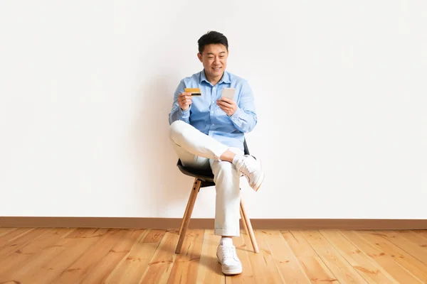 stock image Cheerful asian middle aged man sitting on chair, holding credit card and cellphone, trading on stocks and markets, light wall background, copy space, full length shot