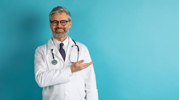 Дружелюбный улыбающийся симпатичный седой врач средних лет в жесте рабочей одежды, показывающий копировальное место для рекламы на синем фоне студии, веб-баннер. Здравоохранение, медицина, концепция клиники