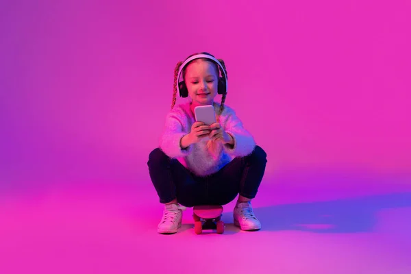 愛らしいかわいいプリティーガールは スケートボードに座って 無線ヘッドフォンを使用して 携帯電話を保持し 友人とチャットし ソーシャルメディアでスクロールし 明るい背景 幼少期の概念 — ストック写真