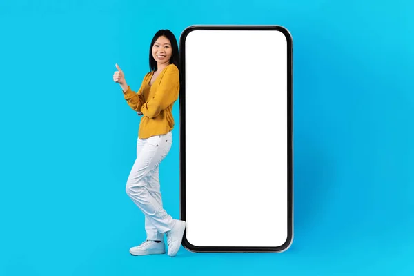 不错的手机应用约会 美丽而快乐的亚洲年轻女子穿着休闲装 被蓝色工作室背景的白色空白屏幕巨大智能手机毒化 露出大拇指 装模作样 — 图库照片