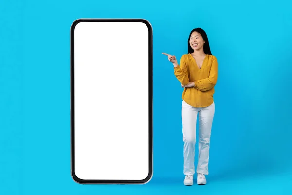 快乐的亚洲年轻女子穿着休闲装 指着蓝色工作室背景上白色空白屏幕的巨大智能手机 用于减肥的移动应用程序 — 图库照片
