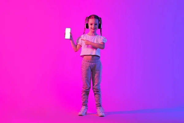 子供のためのビデオコンテンツを楽しんでかなり陽気なスタイリッシュな十代の女の子 オンライン提供のための白い空白の画面のモックアップと携帯電話を示す 無線ヘッドフォンを使用して カラフルな背景 コピースペース — ストック写真