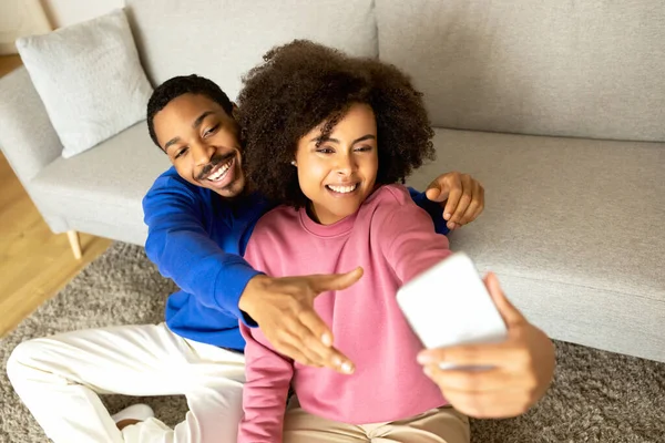 写真好き 楽しいアフリカ系アメリカ人のカップルは 電話でセルフィーを作ります 週末にソーシャルメディアのための幸せな家庭の瞬間をキャプチャ 現代のリビングルームに座ってポーズ 選択的フォーカス — ストック写真