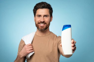 Mutlu orta yaşlı sakallı adam kameraya boş şampuan şişesi gösteriyor ve gülümsüyor, mavi arka planda duruyor, maket. Saç bakım ve vücut bakımı kozmetik konsepti