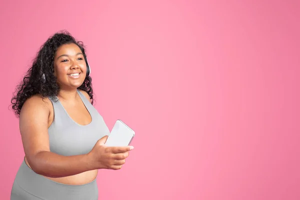 音楽との心臓トレーニング 笑顔黒プラスサイズの女性は ワイヤレスヘッドフォンを身に着けているとピンクの背景にワークアウト中に携帯電話を保持 フリースペース — ストック写真