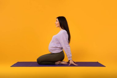 Büyük göbekli, sakin genç bir bayanın egzersiz yaparken, sarı arka planda yoga yaparken yan görüntüsü. Vücut ve sağlık hizmetleri, spor ve hamile çocuk