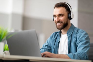 Modern ofiste bilgisayar başında çalışan, kablosuz kulaklık takan, gülümseyen erkek çalışan, masamda oturan ve dizüstü bilgisayarda yazan yakışıklı genç iş adamı, çevrimiçi işten zevk alan, yakın plan