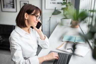 Kahve dükkanında çalışan mutlu kadın iş danışmanı, masada tek başına oturan, dizüstü bilgisayar kullanan ve müşteriyle telefon görüşmesi yapan bir kadın.