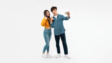 Gadget 'ları severiz. Stüdyoda beyaz arka planda poz veren akıllı telefondan selfie çeken mutlu genç çift. Gençlik Reklam ve Onaylama Mobil Uygulamaları Teklifi. Panorama