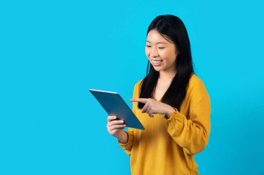 Sarı tişörtlü mutlu Asyalı bayan yepyeni dijital tabletinin tadını çıkarıyor, ekranına dokunuyor, mavi stüdyo arka planında izole edilmiş, kayıyor, güzel bir uygulama kullanıyor, reklam için kopyalama alanı kullanıyor.