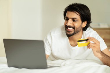 Milenyum ortadoğulu bir adam kredi kartı ve dizüstü bilgisayarla yatakta yatıyor, günaydın, yatak odasının içinde online alışveriş yapıyor. Sipariş ver, nakde çevir, sat, reklam ver ve teklif et.