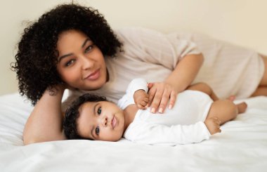 Minik bebeği beyaz yatağa uzanmış neşeli, sevimli siyah milenyum annesi. Aşkın ve yatak odasında boş zamanın tadını çıkarıyor. Güvenli, sevgi, çocuk bakımı, ebeveynlik ve aile evde, reklam ve teklif