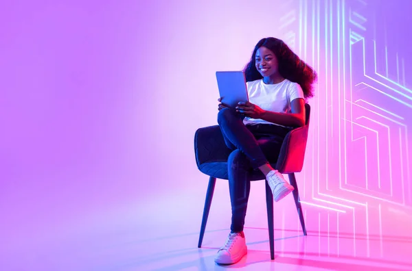 Online life concept. Young positive african american woman web surfing on digital tablet, sitting in armchair over neon light with internet data network, empty space