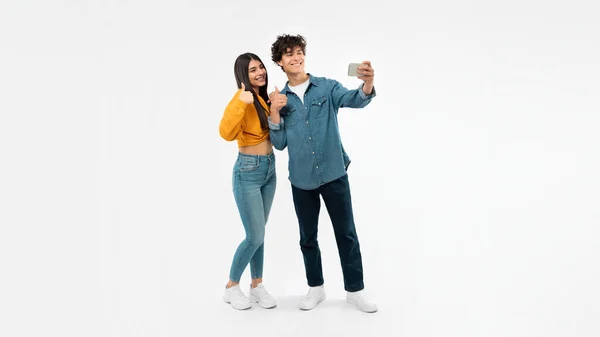 ガジェットが好き スタジオで白い背景に立って立ち上がるスマートフォンのジェスチャー親指でセルフィーを作る幸せな若いカップル 若者の広告と承認モバイルアプリの提供 パノラマ — ストック写真