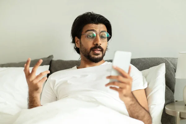 悲喜交集的中东千禧年男人戴着眼镜 在电话上看到消息 躺在床上 躺在卧室的内部 坏消息 远程沟通 工作中的问题 广告和提供 — 图库照片