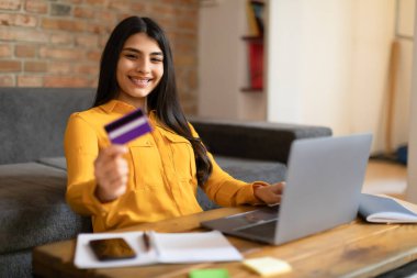 Neşeli İspanyol bayan evde online alışveriş yapıyor, kredi kartı ve dizüstü bilgisayar kullanıyor, internetten bir şeyler sipariş ediyor, oturma odasında otururken bir şeyler alıyor, yer kopyalıyor.