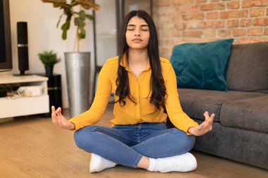 Sakin İspanyol bayan kapalı gözlerle meditasyon yapıyor, evde oturma odasında yerde oturuyor, huzurlu kadın yoga yapıyor, içsel dengeyi buluyor, stres yönetiyor.