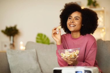 Dijital Eğlence. Neşeli Afro-Amerikan Kadın Projektörü Sevdiği Filmi Kullanıyor, Modern Oturma Odasında Patlamış Mısır Yiyiyor, Evdeki Koltukta Eğleniyor. Hafta sonu Eğlencesi