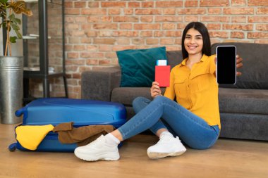 Neşeli genç İspanyol bayan turist valizinin yanında oturuyor, elinde uçuş biletleriyle pasaportunu tutuyor ve cep telefonunu boş ekran ile gösteriyor, uygulama veya web sitesi için maket yapıyor.