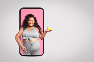 Gri stüdyo arka planında izole edilmiş akıllı telefon ekranında pulları ve elması olan, spor giyim sektöründe sırıtan, iri yarı bir kadın. Vücut bakımı, spor, kilo kaybı ve zindelik uygulaması