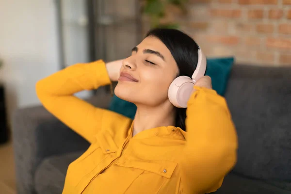 静かな若いスペイン人女性は ワイヤレスヘッドフォンで音楽を聞いて 目を閉じて 自宅でソファに座って休んで頭の後ろに手を保持ソファにリラックス — ストック写真
