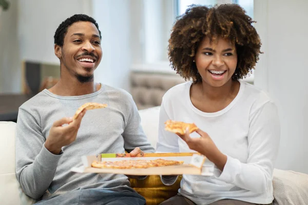 幸せな若い黒のカップルの家庭では コピースペースと笑顔を見てピザを食べている リラックスしたアフリカ系アメリカ人の男性と女性は ソファに座っているパジャマを着て 週末を楽しんで 映画を見て — ストック写真