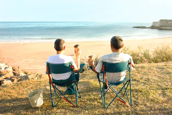 海洋周末 资深旅行家们坐在椅子上回到相机前 喝着纸杯里的咖啡 看着外面的海滩和海水 配偶度假放松的后视镜 — 图库照片