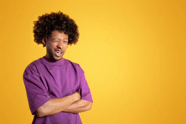 Positive confident mature black curly guy in purple t-shirt with crossed arms on chest, winks eye isolated on yellow background, studio. Lifestyle, hint for sale, recommendation, ad and offer