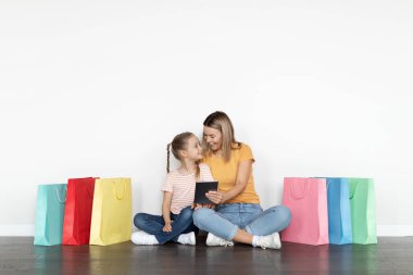 Alışveriş Uygulaması. Neşeli genç bir anne ve dijital tabletli küçük bir kız. Parlak alışveriş torbalarının arasında oturuyorlar. Mutlu anne ve çocuklar çevrimiçi alışverişlerin ve sezonluk satışların keyfini çıkarıyorlar.