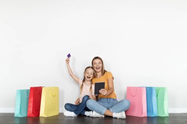 Kolay alışveriş. Heyecanlı küçük kız ve mutlu annesi internet alışverişi için dijital tablet ve kredi kartı kullanıyor. Parlak kağıt torbaların arasında oturuyor, online satışların keyfini çıkarıyor.