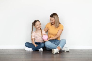 Aile Birikimleri. Mutlu genç anne ve şirin küçük kız domuz kumbarasını tutarken beyaz duvarın yanında dinleniyor, annesine para biriktirmeyi ve ekonomiyi öğretiyor,