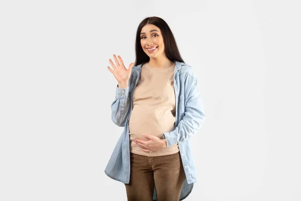 积极的年轻孕妇摸着肚子 在镜头前挥手打招呼和微笑 摆出一副白色工作室的样子 友好的欢迎姿态 — 图库照片