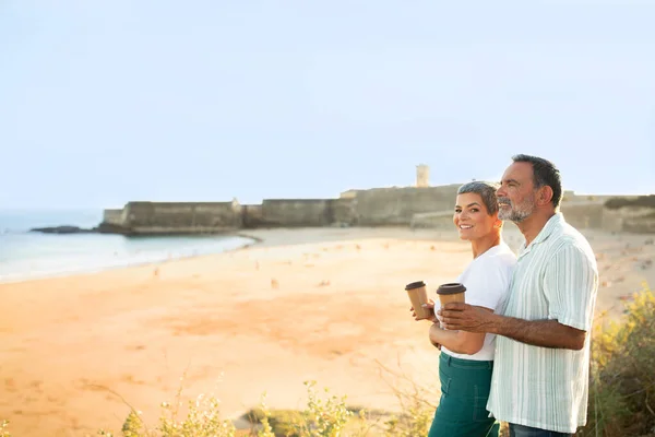 快乐的老年旅伴们 带着咖啡杯来到海洋的户外 喝着热饮着 在海滨享受着这一天 爱的配偶的侧视图 在近海时拍摄 文字的自由空间 — 图库照片