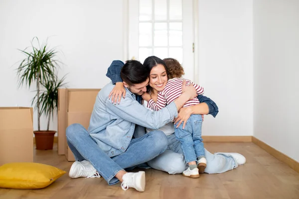 Taşınma Günü Kutlaması. Mutlu bir aile, yeni bir evde kucaklaşan mutlu bir aile, neşeli bir genç ebeveyn ve yeni yürümeye başlayan çocukları, karton kutuların arasında oturuyor, birbirlerine sarılıyor, şefkat dolu anlar yaşıyorlar.