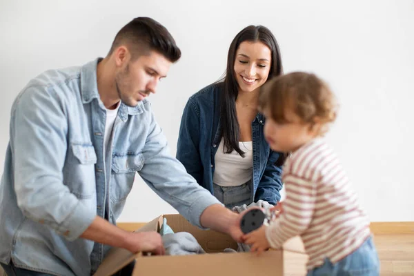 Young Family Of Three Packing Things Together While Moving Home, Parents And Their Cute Toddler Son Putting Belongings Into Cardboard Box, Getting Ready For House Relocation, Selective Focus