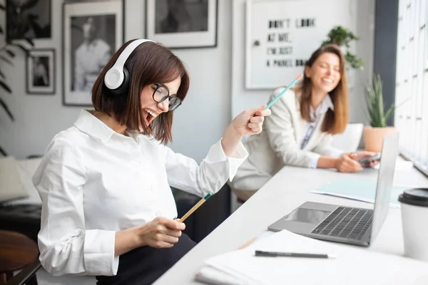 stock image Joyful lady office manager in headphones enjoying music and singing while working on laptop in office, sitting with colleague at workplace, selective focus