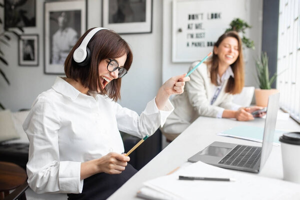 Joyful lady office manager in headphones enjoying music and singing while working on laptop in office, sitting with colleague at workplace, selective focus