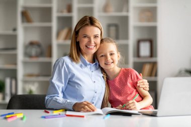 Güzel bir anne ve küçük kızının evde masada oturup kameraya gülümseyip kucaklaşması, mutlu bir anne ve ev ödevi yapan bir kız çocuğunun portresi.