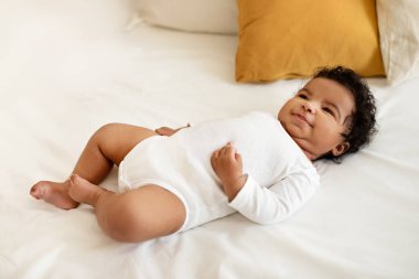 Neşeli, gülümseyen, küçük, siyah, tatlı çocuk, beyaz bir yatakta uzanıyor. Sabah yatak odasında, manzaralı, uzun boylu. Yeni doğan, çocukluk, çocuk bakımı, evde ebeveynlik, reklam ve sunum