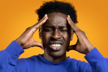 Siyah adam baş ağrısı ya da migren hissediyor, şakaklara masaj yapıyor, sarı arka planda aşırı çalışmaktan yorgun düşmüş. Gürültülü ses, baş dönmesi, baş ağrısı ve yakın çekim yüzünden bunalmış genç bir adam.