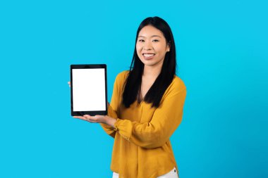 En yeni mobil uygulama. Mutlu, güzel, uzun saçlı, Asyalı bir kadın beyaz ekranlı, gülümseyen, mavi stüdyo arka planında izole edilmiş yepyeni dijital tablet gösteriyor.